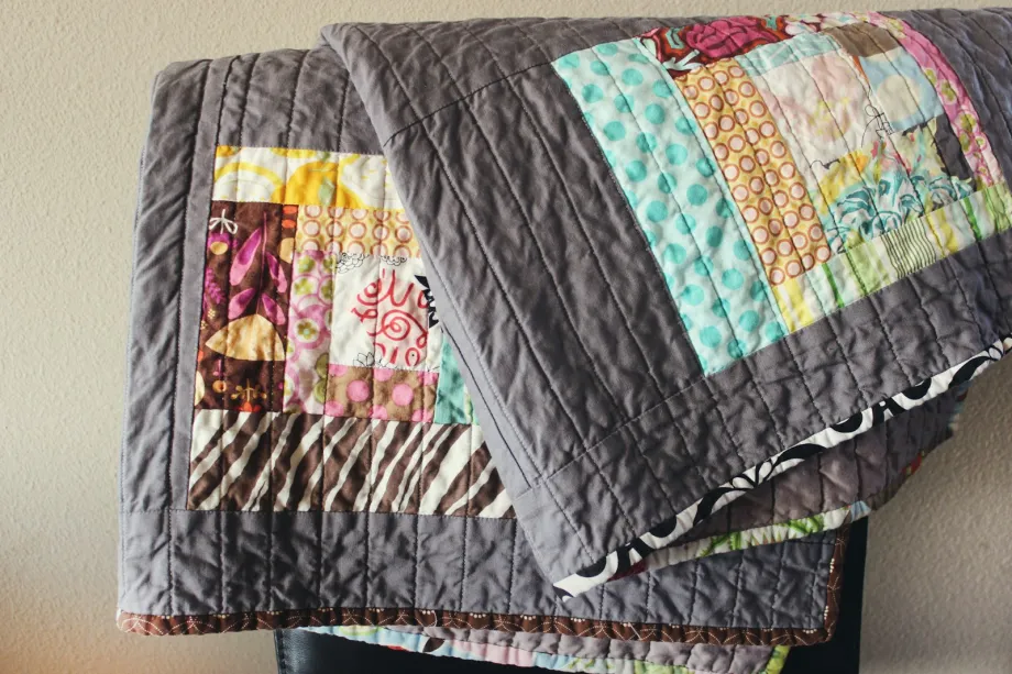 Quilt with blocks of colorful print strips set in gray, with straight-line quilting, hung over a chair