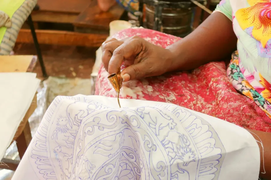 Hand applying wax with a small metal spouted tool to cloth with a drawn batik design